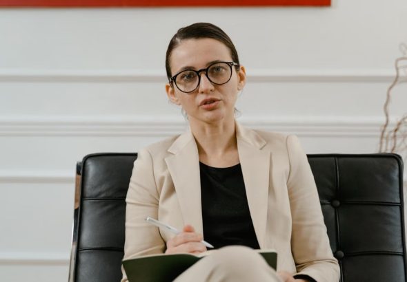 A woman in a blazer writing notes on a clipboard while sitting in an office.