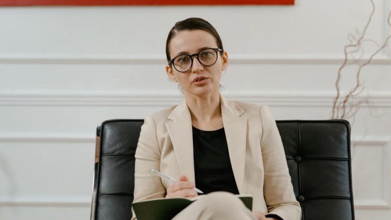 A woman in a blazer writing notes on a clipboard while sitting in an office.